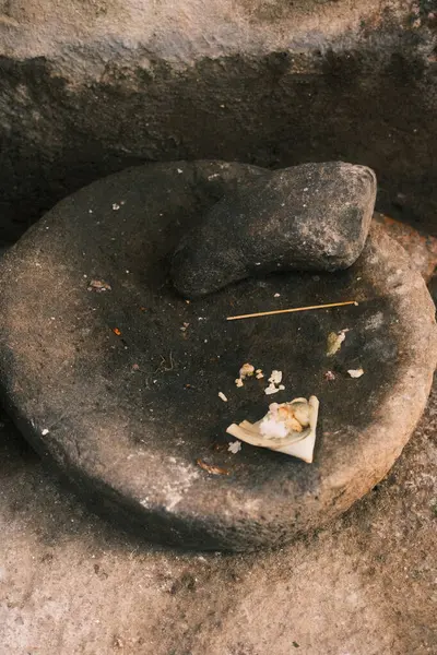 Bu resim yıpranmış, geleneksel bir taş havan ve tokmak yakalar, beyaz adak malzemesi kalıntıları ile, sert, topraksı bir yüzeye yerleşir, Balili günlük yaşamın tipik bir örneği..