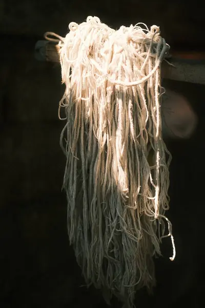 Bu resim karmaşık bir demet açık renkli, muhtemelen beyaz ya da bej, lifler ya da ipler içeriyor, eski bir paspas başı gibi görünüyor, koyu bir arkaplanda asılı, bir güneş ışığı kuyusuyla aydınlatılıyor..
