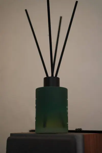 Siyah kamışlı mat yeşil cam kamış yayıcı, düz bir ışık duvarına karşı karanlık bir yüzey üzerinde duruyor, minimalist ve sakin bir atmosfer yaratıyor.