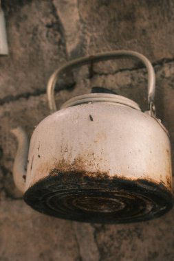 Sert, desenli tuğla bir duvara dayanmış ağır yanmış ve paslı tabanı olan eski, beyaz metal bir su ısıtıcısının yakın çekimi, kırsal ya da geleneksel bir mutfak düzenini işaret ediyor.