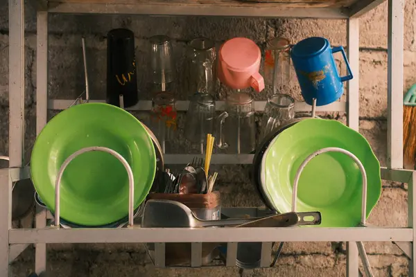 Parlak yeşil tabaklar, bardaklar, bardaklar ve kırsal taştan bir duvara dayanan çatal bıçaklar da dahil olmak üzere çeşitli mutfak malzemeleriyle dolu çok katmanlı metal tabak rafı.,