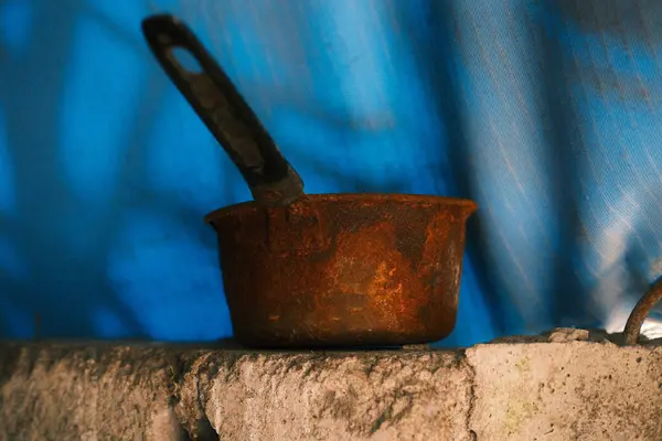 Ağır paslanmış, uzun saplı eski metal tencereye yakın plan, pürüzlü, düz olmayan bir taş ya da beton yüzey üzerinde duruyor, bulanık mavi ve beyaz arka plan üzerine kurulmuş.