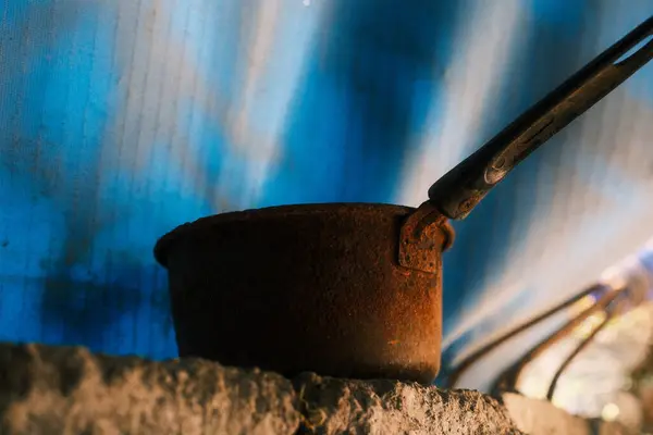 Ağır paslanmış, uzun saplı eski metal tencereye yakın plan, pürüzlü, düz olmayan bir taş ya da beton yüzey üzerinde duruyor, bulanık mavi ve beyaz arka plan üzerine kurulmuş.