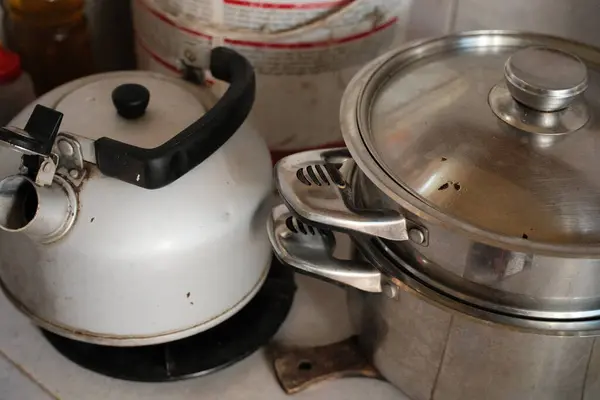 Bu resim, eski, beyaz metal bir su ısıtıcısını ve kapaklı paslanmaz çelik bir tencereyi yakalar. Kullanım ve yaşlanma belirtilerini gösterir, dokulu bir yüzeyde dinlenir.