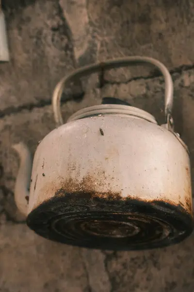 Sert, desenli tuğla bir duvara dayanmış ağır yanmış ve paslı tabanı olan eski, beyaz metal bir su ısıtıcısının yakın çekimi, kırsal ya da geleneksel bir mutfak düzenini işaret ediyor.