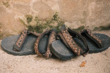 Bir çift iyi kullanılmış siyah sandalet belirgin desenli kayışlar beton bir yüzeye dayanır, yıpranmış bir duvarla desteklenir ve yıpranma izleri gösterir..