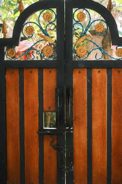 Büyüleyici bir bahçe kapısına yakın plan, koyu işlemeli demirle karışık altın çiçekli motifleri ve sıcak ahşap panelleri harmanlıyor. Ev dekorasyonu, güvenlik ve geleneksel zanaat temaları için ideal