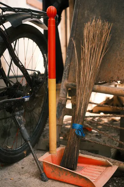 Kırmızı ve sarı saplı geleneksel bir süpürge bisiklet tekerinin yanındaki turuncu bir faraş üzerinde duruyor. Bu da günlük ev işlerini ya da etrafı toplamaya ara vermeyi öneriyor..