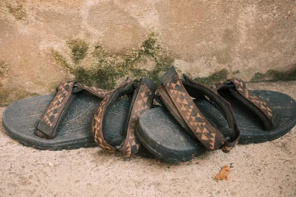 Bir çift iyi kullanılmış siyah sandalet belirgin desenli kayışlar beton bir yüzeye dayanır, yıpranmış bir duvarla desteklenir ve yıpranma izleri gösterir..