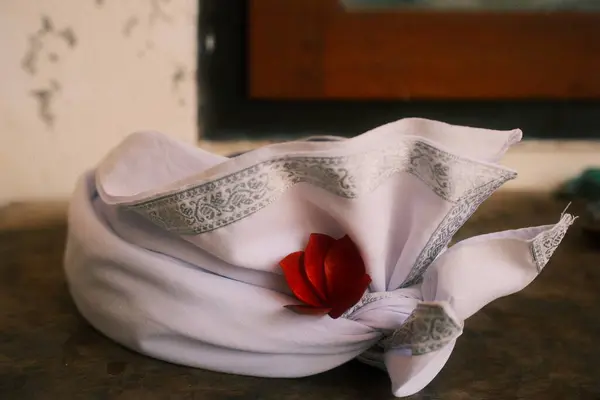 Güzelce katlanmış beyaz bir Balili Udeng, geleneksel bir başörtüsü, karmaşık gümüş desenli ve canlı bir kırmızı çiçekle süslenmiş, dokulu bir yüzeye dayanır, Bali kültürünü ve ruhaniliğini sembolize eder..