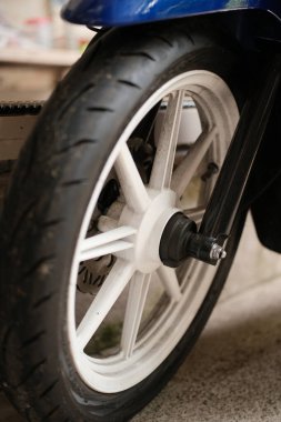 Yakın çekim motorsiklet ya da scooter 'ın ön tekerleğine odaklanır, beyaz jantlarını, lastik izli siyah lastiğini ve beton kaldırımda dinlenen mavi çamurluğun bir kısmını vurgular..