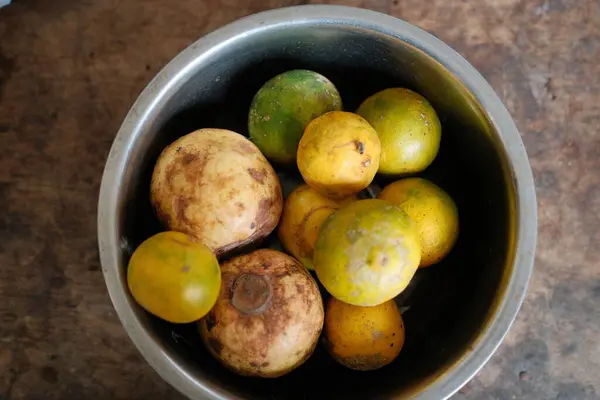 Tepeden çekimde taze, olgun ve olgunlaşmamış yeşil limonlar, sarımsı mandalinalar ve soluk yeşil guavalar içeren çeşitli tropikal meyvelerle dolu bir metal kâse sergileniyor.