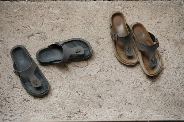 Denpasar, Bali - 7 Mayıs 2025: İki çift sıradan erkek parmak arası terlik, bir koyu gri ve açık kahverengi, desenli beton bir yüzey üzerinde dinleniyor, rahat ve rahat bir günlük ayakkabı titreşimini çağrıştırıyor açık ya da rahat bir ortam için