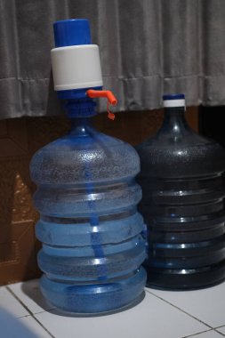 İki büyük plastik içme suyu galonu, biri mavi ve beyaz bir pompa dağıtıcısı ile donatılmış, arka planda bulanık perdelerle döşenmiş, ev hidrasyonunu temsil eden.