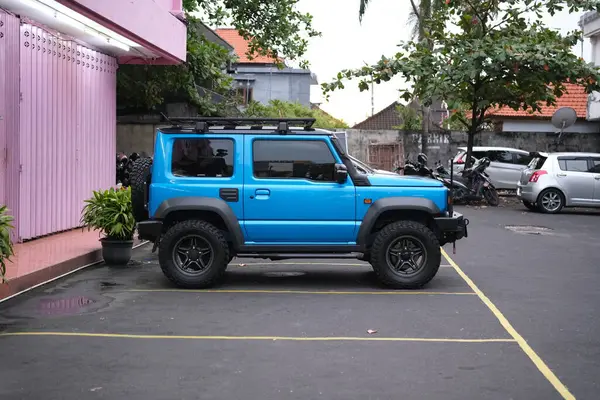 Çatı rafı ve büyük lastiklerle donatılmış canlı mavi bir SUV, pembe bir bina ve diğer araçların bulanık arka planına kurulmuş sarı işaretli bir asfalt alana park edilmiştir.