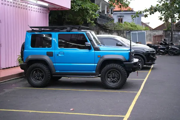 Çatı rafı ve büyük lastiklerle donatılmış canlı mavi bir SUV, pembe bir bina ve diğer araçların bulanık arka planına kurulmuş sarı işaretli bir asfalt alana park edilmiştir.