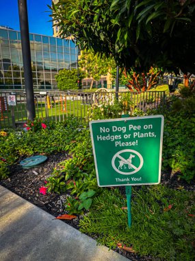Los Angeles, Kaliforniya, ABD - 25 Mayıs 2025: Çitlere Köpek Çişi Yok ya da Bitkiler Lütfen İmzala, Yeşil ve Beyaz Metal Üstü Çapraz Köpek, Pembe Çiçekler, Ağaçlar ve Çalılıklar, Arka planda Los Angeles Skyline