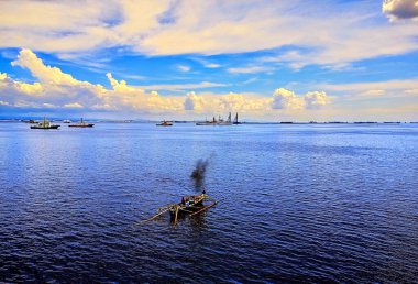 Geleneksel Filipinli bangka teknesi okyanusun ortasında, küçük ve ahşap, arka planda büyük gemiler görünüyor..