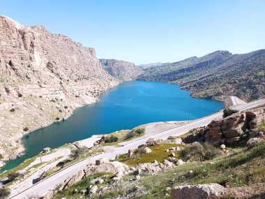 Geniş açı, engebeli, kayalık dağların arasına kurulmuş büyük, canlı bir mavi gölü gösteriyor. Gölün kıyısında bir yol esiyor ve baraj yapısının bir kısmı sağda görünüyor. Yeşil bitki örtüsü dağların alçak yamaçlarında yer alır..