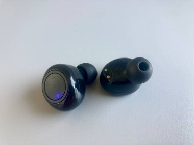 Bir çift parlak siyah kablosuz kulaklık beyaz bir yüzeyde duruyor. Bir tanesi, minimalist tasarım ve şarj konnektörleri görünür durumda, güç veya bluetooth modu öneren parlayan mavi bir LED ışığı içerir.