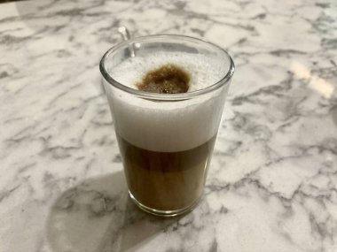 İnce köpüklü şeffaf bir bardak kahve rafine mermer bir yüzeye oturur. Zengin kahve tonları ve yumuşak köpük, ışıl ışıl tezgahla uyumlu bir şekilde harmanlanarak sıcaklığı ve karmaşıklığı çağrıştırıyor..