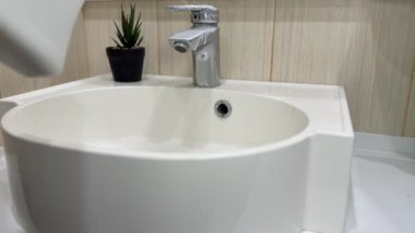 Bir adam güçlü bir sprey temizleyici kullanarak banyo lavabosunu temizliyor. Sarı lastik eldivenler giyerek musluğu ve yüzeyleri süngerle temizliyor..