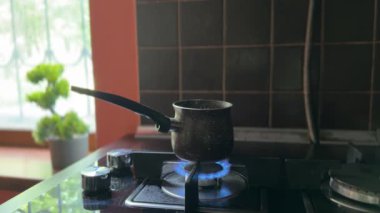 Loş bir ev mutfağındaki gaz sobasının üzerinde kahverengi bir kahve duruyor. Sahne, kahve kaynamaya başlamadan önceki sakin anı yakalar, samimi titreşimler uyandırır..