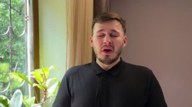Alerji krizinden muzdarip bir adam dairesinde duruyor. Keskin bir şekilde hapşırır ve burnunu silmek için mendil kullanır. Alerjik rhinitis belirtileri görülebilir..