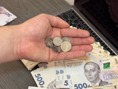 Birkaç bozuk para, arka planda para ve dizüstü bilgisayarla açık avuçta duruyor. Görüntü, çevrimiçi faaliyetlerden elde edilen finans, tasarruf veya dijital geliri sembolize eder.