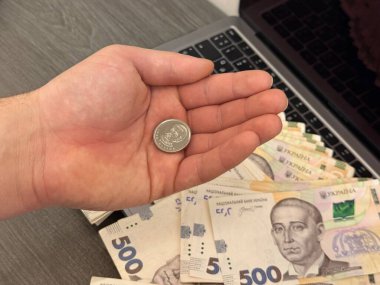 Tek bir madeni para, yoksulluğu temsil eden açık bir avuç içinde dururken, bir dizüstü bilgisayar ve 500 UAH banknot yığını arka planda oturup zenginliğin karşıtlığını gözler önüne seriyor..