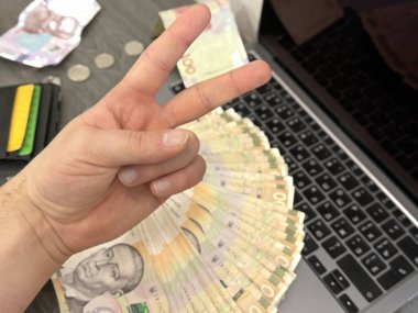 Cüzdanı, bilgisayarı ve 500 UAH banknotuyla bir masanın önündeki zafer işaretini gösteren bir el. Başarı, para ve dijital çalışmanın sembolü.