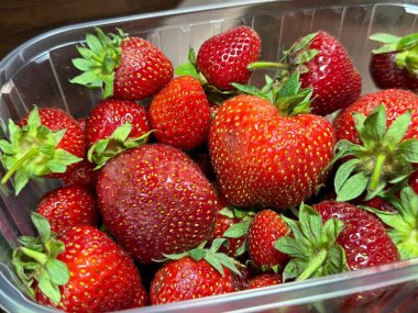 Çilekleri karanlık bir masada plastik bir kapta yakın plan çekiyoruz. Bazı meyveler bozulma belirtileri gösteriyor. Fotoğrafta gıda tazeliği ve depolama sorunları vurgulanıyor.