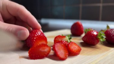 Tahta bir tahtanın üzerinde olgun çilekleri kesmeye yakın çekim. Bir çilek dilimlenmiş, diğerleri bekliyor. Sulu meyveli yaz mutfağı hazırlık sahnesi.