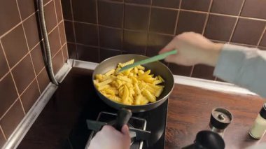 Patates kızartmasını bir tavada kızartırken bir kişi de onları spatulayla karıştırıyor. Sıcak altın patatesler mutfak sobasında pişiyor, lezzetli ev yapımı fast food gösteriyor..