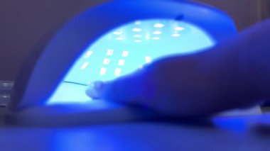 Genç bir kadın, bir ultraviyole lambanın manikür sırasında jel ojeleri iyileştirmek için nasıl kullanıldığını gösteriyor. Süreç, tırnak bakımı, güzellik rutini ve modern teknikleri vurguluyor..