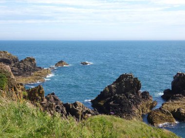 Yumuşak, hafif bulutlu bir gökyüzünün altında sakin mavi denize dalan pürüzsüz, siyah kayalıklı bir kıyı şeridi. Küçük su birikintileri kaya oluşumları arasında dinleniyor, hafif dalgalar kenarlara vuruyor..