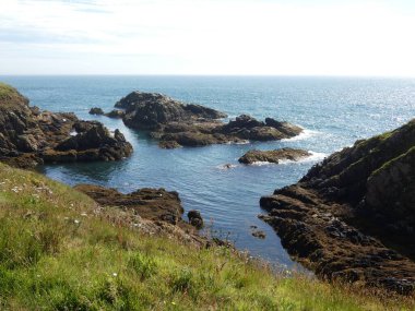 Yumuşak, hafif bulutlu bir gökyüzünün altında sakin mavi denize dalan pürüzsüz, siyah kayalıklı bir kıyı şeridi. Küçük su birikintileri kaya oluşumları arasında dinleniyor, hafif dalgalar kenarlara vuruyor.. 