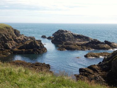 Yumuşak, hafif bulutlu bir gökyüzünün altında sakin mavi denize dalan pürüzsüz, siyah kayalıklı bir kıyı şeridi. Küçük su birikintileri kaya oluşumları arasında dinleniyor, hafif dalgalar kenarlara vuruyor.. 