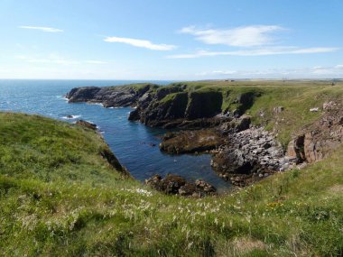 Yumuşak, hafif bulutlu bir gökyüzünün altında sakin mavi denize dalan pürüzsüz, siyah kayalıklı bir kıyı şeridi. Küçük su birikintileri kaya oluşumları arasında dinleniyor, hafif dalgalar kenarlara vuruyor..