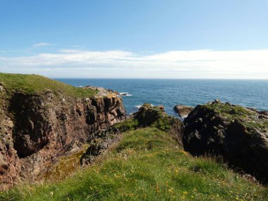 Yumuşak, hafif bulutlu bir gökyüzünün altında sakin mavi denize dalan pürüzsüz, siyah kayalıklı bir kıyı şeridi. Küçük su birikintileri kaya oluşumları arasında dinleniyor, hafif dalgalar kenarlara vuruyor.. 