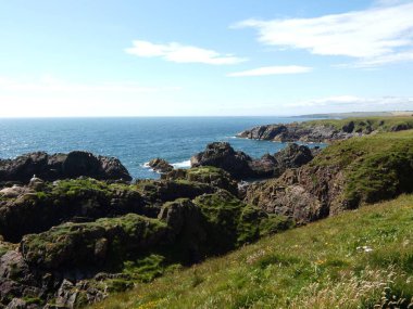 Yumuşak, hafif bulutlu bir gökyüzünün altında sakin mavi denize dalan pürüzsüz, siyah kayalıklı bir kıyı şeridi. Küçük su birikintileri kaya oluşumları arasında dinleniyor, hafif dalgalar kenarlara vuruyor.. 