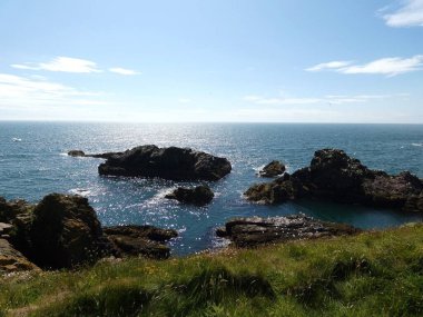 Yumuşak, hafif bulutlu bir gökyüzünün altında sakin mavi denize dalan pürüzsüz, siyah kayalıklı bir kıyı şeridi. Küçük su birikintileri kaya oluşumları arasında dinleniyor, hafif dalgalar kenarlara vuruyor.. 