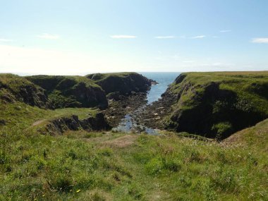 Yumuşak, hafif bulutlu bir gökyüzünün altında sakin mavi denize dalan pürüzsüz, siyah kayalıklı bir kıyı şeridi. Küçük su birikintileri kaya oluşumları arasında dinleniyor, hafif dalgalar kenarlara vuruyor.