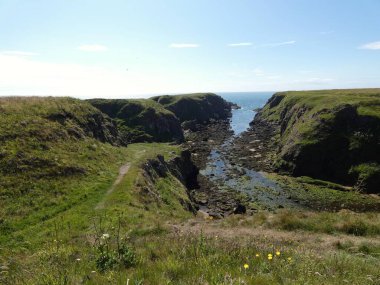 Yumuşak, hafif bulutlu bir gökyüzünün altında sakin mavi denize dalan pürüzsüz, siyah kayalıklı bir kıyı şeridi. Küçük su birikintileri kaya oluşumları arasında dinleniyor, hafif dalgalar kenarlara vuruyor.. 