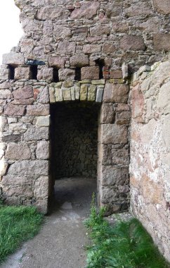 Kaba yontulmuş granit bloklar tarafından çerçevelenmiş eski bir kalenin kalın duvarlarına yerleştirilmiş yıpranmış taş bir kapı. Dar kemerli giriş karanlık, kapalı bir taş odaya açılıyor. İçi ancak doğal ışıkla aydınlanıyor..