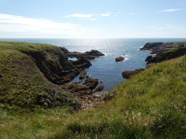 Yumuşak, hafif bulutlu bir gökyüzünün altında sakin mavi denize dalan pürüzsüz, siyah kayalıklı bir kıyı şeridi. Küçük su birikintileri kaya oluşumları arasında dinleniyor, hafif dalgalar kenarlara vuruyor.. 