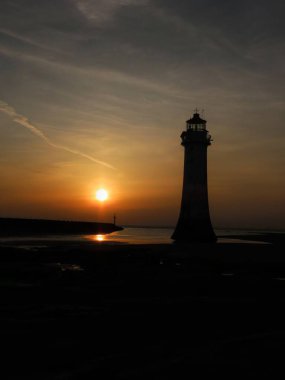 Ufukta New Brighton Deniz Feneri nefes kesici bir günbatımına karşı muhteşem bir şekilde duruyor. Gökyüzü ılık turuncu ve sarı renklerle parıldıyor, durgun sulara ve çevreleyen kıyı şeridine dingin bir ışık saçıyor..