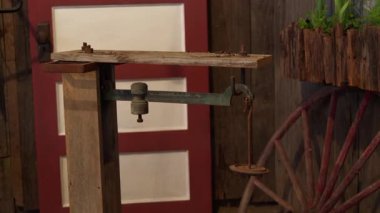 Metal kol ve ağırlıkla eski usul mekanik denge terazisi, ahşap bir standa monte edilmiş. Arka planda kırmızı ve beyaz ahşap kapı, bir araba tekerleği ve yeşil bitkilerle dolu bir saksı..