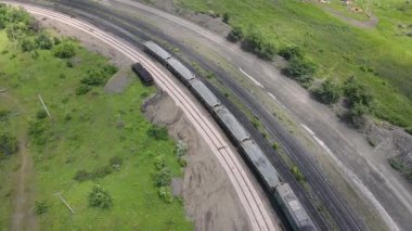 Yeşil tarlalar ve toprak yollarla çevrili bir tren yolunda keskin bir dönüş yapan bir yük treninin hava aracı görüntüsü, lokomotiften gelen dumanla. 