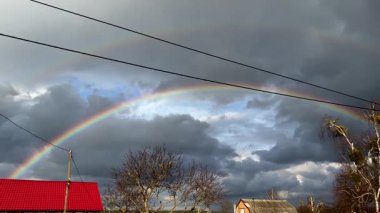 Karanlık, kasvetli gökyüzünde canlı bir gökkuşağı beliriyor. Aşağıda, yapraksız ağaç siluetleri ve elektrik hatları var. Bu atmosferik açık hava sahnesinde dramatik bir kontrastlık.